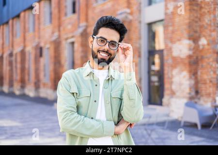 Porträt eines jungen Mannes in lässiger Kleidung, fröhlicher Geschäftsmann, der lächelnd und mit gekreuzten Armen in die Kamera blickt, indischer Mann vor dem Bürogebäude. Stockfoto