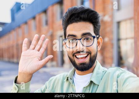 Webkamera-Ansicht des Telefons, junger Mann verwendet App auf dem Smartphone für die Fernkommunikation von Videoanrufen, indischer Mann lächelt und schaut in die Kamera, winkt Hand in Begrüßung. Stockfoto