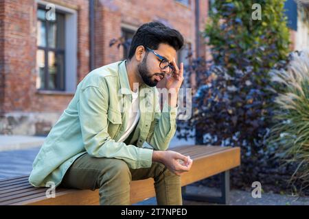 Kranker Mann, der auf einer Bank vor dem Bürogebäude sitzt, indischer Mann mit schweren Kopfschmerzen und Schwindel Migräne, überarbeiteter und überarbeiteter Arbeiter in legeren Kleidern. Stockfoto