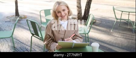 Bild einer jungen lächelnden Frau, die in einem Café im Freien sitzt, Skizzen macht, Kunst auf der Straße zeichnet, Zeit im Café verbringt und Kunst kreiert Stockfoto