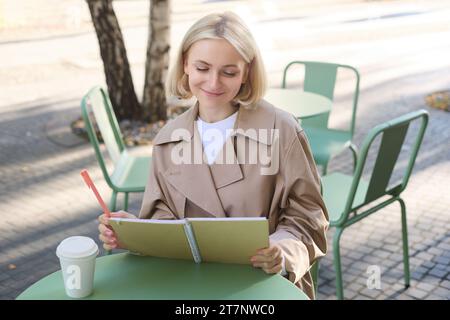 Porträt einer jungen kreativen Frau, die im Freien skizziert, mit Notizbuch und Stift im Café sitzt, Kunst zeichnet und lächelt Stockfoto