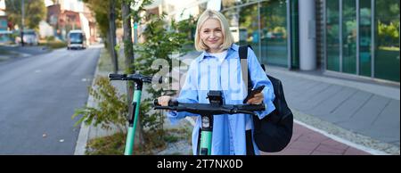 Frau scannt QR-Code am Lenker eines gemeinsam genutzten Elektrorollers. Nahaufnahme von weiblicher Hand, die das Smartphone hält, um den grünen E-Scooter in der Straße freizuschalten Stockfoto