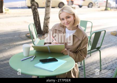 Bild einer jungen lächelnden Frau, die in einem Café im Freien sitzt, Skizzen macht, Kunst auf der Straße zeichnet, Zeit im Café verbringt und Kunst kreiert Stockfoto