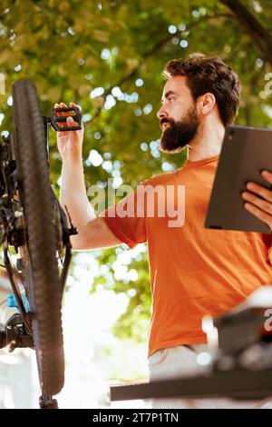 Männlicher kaukasischer Radfahrer, der Fahrrad gründlich mit einem digitalen Tablet inspiziert und repariert. Detaillierte Aufnahme eines gesunden sportliebenden Mannes auf der Suche nach Fahrrad-Sommer-Wartung auf dem Handy-Tablet. Stockfoto