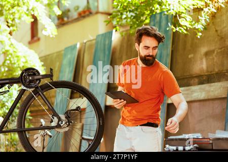 Energischer Radfahrer, der sein digitales Tablet greift, während er nach wichtigen Arbeitsgeräten für die Inspektion und Reparatur von Fahrrädern im Hof sucht. Aktiver kaukasischer Mann, der sich auf die Sommerwartung vorbereitet. Stockfoto