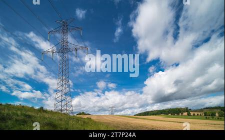 Oberleitungsmast in der Sommerlandschaft. Elektrischer Traversen-Übertragungsturm mit Hochspannungsdrähten, grünem Gras, Stoppeln, Feld oder Bäumen am Berg. Stockfoto