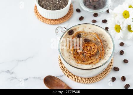 Chia-Samenpudding mit Kaffee und Erdnussbutter in Glas, Holzlöffel und Blumen auf weißem Marmortisch. Kopierbereich. Gesundes Frühstücksgetränk. Stockfoto