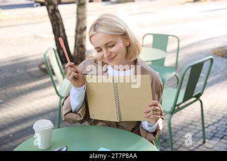 Porträt einer jungen kreativen Frau, die im Freien skizziert, mit Notizbuch und Stift im Café sitzt, Kunst zeichnet und lächelt Stockfoto