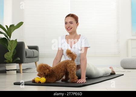 Glückliche junge Frau, die Yoga auf Matte mit ihrem süßen Hund zu Hause praktiziert Stockfoto