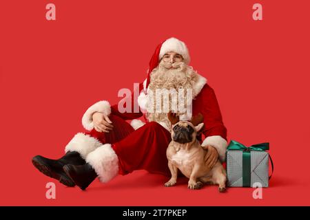 Weihnachtsmann mit Weihnachtsgeschenk und französischer Bulldogge in Rentierhörnern auf rotem Hintergrund Stockfoto