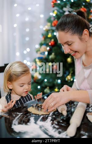 Mom legt Keksschneider auf den Teig in der Nähe ihrer kleinen Tochter Stockfoto