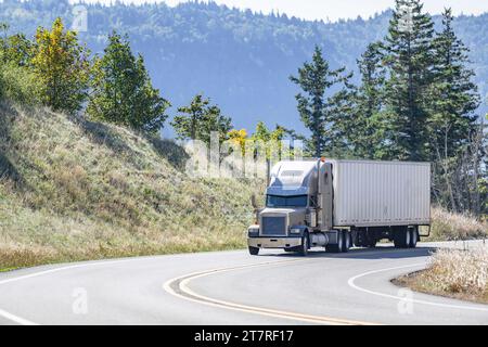 Industrieller beigefarbener klassischer Carrier großer Lkw, der gewerbliche Güter in einem trockenen Anhänger transportiert und auf der kurvenreichen Bergstraße bergauf fährt Stockfoto