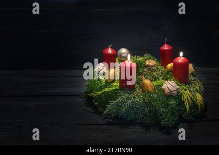 Licht im Dunkeln am zweiten Advent, naturgrüner Kranz mit roten Kerzen, zwei brennen, Weihnachtsdekoration und Kekse, dunkler hölzerner Hintergrund, Stockfoto