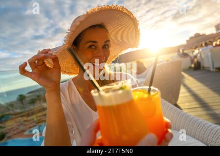 Frau im Sonnenhut lächelt und toast mit einem Cocktail im tropischen Strandhotel bei Sonnenuntergang mit verschwommenem Hintergrund Stockfoto