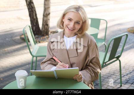 Bild einer jungen lächelnden Frau, die in einem Café im Freien sitzt, Skizzen macht, Kunst auf der Straße zeichnet, Zeit im Café verbringt und Kunst kreiert Stockfoto