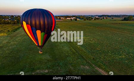 Ein Luftblick auf einen schwarz gestreiften Heißluftballon, der an einem sonnigen Sommermorgen tief über Felder und Ackerland im ländlichen Amerika schwimmt. Stockfoto