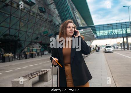 Geschäftsfrau, die sich vom Flughafen beeilt, auf der Straße der Stadt steht und einen Pass mit Tickets hält und einen Koffer bei sich trägt, während sie ein Taxi per Handy anruft Stockfoto