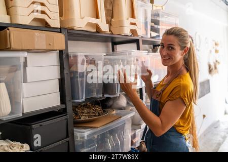 Eine Frau in einem senffarbenen Hemd und einer Denim-Schürze stellt in einer Werkstatt für getrocknete Blumen klare Aufbewahrungsbehälter auf Regalen und lächelt, während sie sich anschaut Stockfoto