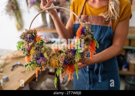 Florist in einer blauen Schürze mit einem bunten und lebendigen getrockneten Blumenkranz in einer Werkstatt Stockfoto