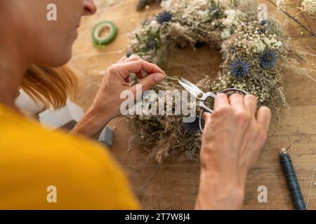 Nahaufnahme der Hände eines Floristen mit einer Schere, um einen getrockneten Blumenkranz auf einem Holztisch zu bearbeiten Stockfoto