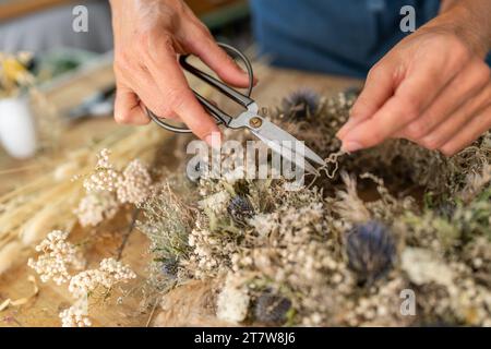 Hände mit einer Schere einen getrockneten Blumenkranz auf einem hölzernen Werkstatttisch zuschneiden Stockfoto