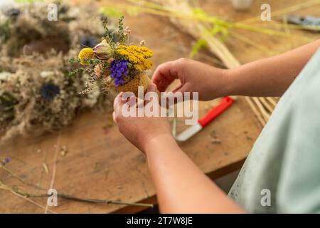 Hände, die einen kleinen Strauß bunter getrockneter Blumen in einem Kegel auf einem Holztisch basteln Stockfoto