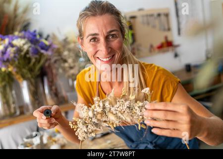 Lächelnde Floristin in einer gelben Spitze, die ein getrocknetes Blumenarrangement in einer Werkstatt schafft Stockfoto