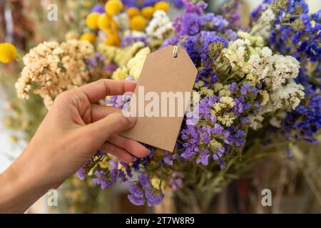 Halten Sie ein leeres braunes Etikett vor einem Strauß aus bunten getrockneten Blumen, mit Copyspace für Ihren individuellen Text. Stockfoto