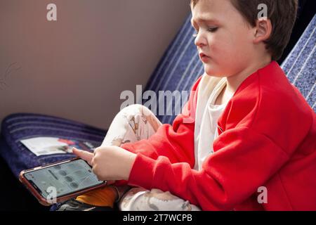 Tech-Savvy Journey: Ein 6-jähriger Junge, der während einer Zugfahrt in seine Smartphone-Spiele vertieft ist, sein engelhaftes Gesicht voller Konzentration. Stockfoto