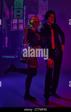 Frau in Schuluniform mit Siegeszeichen in der Nähe des Mannes auf lila Neonhintergrund mit Hieroglyphen Stockfoto