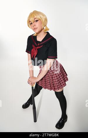 Die ganze Länge einer Anime-Frau in trendiger Schuluniform mit Baseballschläger, die in weiß in die Kamera schaut Stockfoto