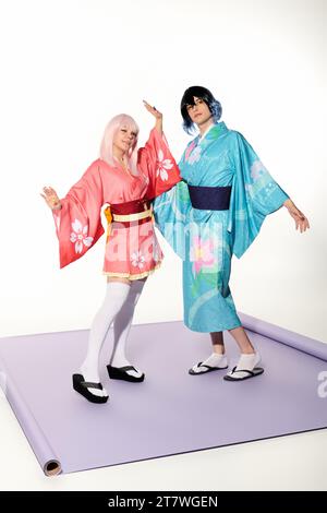 Junge ausdrucksstarke Cosplayer in bunten Kimonos und Perücken, die auf lila Teppich im weißen Studio posieren Stockfoto