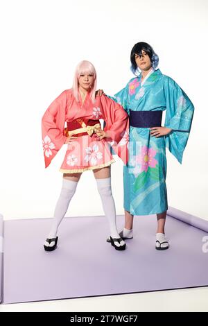 Selbstbewusstes ausdrucksstarkes Paar in bunten Kimonos und Flügeln, das mit Händen auf der Hüfte posiert, Cosplay-Trend Stockfoto