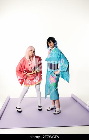 Junge verspielte Cosplayer in Kimonos und Perücken mit Händen auf Hüften auf lila Teppich im weißen Studio Stockfoto