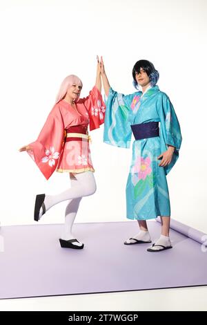 Fröhliche Cosplayer in hellem Kimono und Perücken, die hohe fünf auf lila Teppich und weißem Hintergrund geben Stockfoto