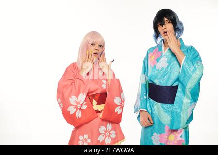 Erstauntes Anime-Paar in bunten Kimonos und Perücken, die Gesichter auf weißem Cosplay-Trend berühren Stockfoto