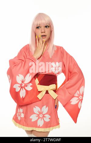 Schockierte Anime-Stil-Frau in Pink mit Hand auf Hüfte berührendes Gesicht auf weißem Cosplay-Charakter Stockfoto