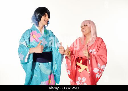 Fröhliche Cosplayer in bunten Kimonos und Perücken, die Daumen nach oben zeigen und sich auf weiß ansehen Stockfoto