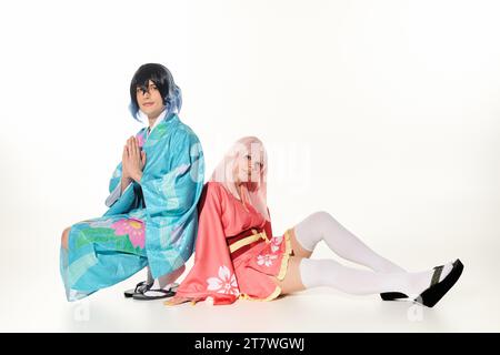 Anime-Stil Mann im Kimono mit betenden Händen in der Nähe der Frau in blonder Perücke, die auf weiß sitzt, Cosplayer Stockfoto