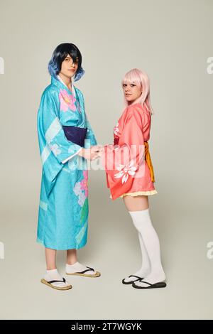 Ein junges Paar in bunten Kimonos, das Hände hält und die Kamera auf grauem Anime-Konzept ansieht Stockfoto