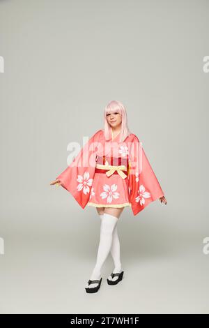 Die ganze Länge einer jungen Anime-Frau in blonder Perücke und rosa Kimono auf grauer, japanischer Subkultur Stockfoto