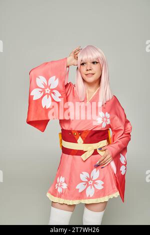 Junge lächelnde Frau in blonder Perücke und rosa Kimono mit Blumenmuster, die auf grauem Anime-Stil schweben Stockfoto