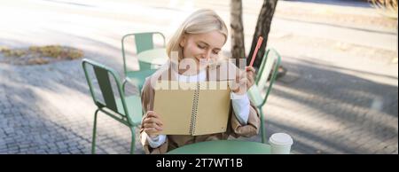 Porträt einer jungen kreativen Frau, die im Freien skizziert, mit Notizbuch und Stift im Café sitzt, Kunst zeichnet und lächelt Stockfoto