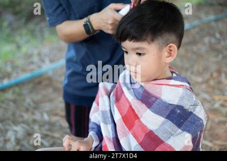 Friseur schneidet Haare eines asiatischen Jungen in einem offenen Raum voller Bäume. Stockfoto