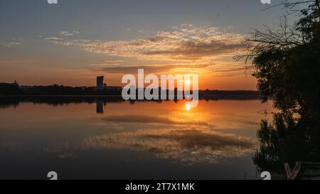 Malerische Sonnenaufgangssonne auf dem See mit bewölktem Himmel, der sich im ruhigen Wasser spiegelt. Friedlicher Morgen am Fluss Stockfoto