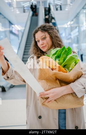 Eine schockierte Frau sieht sich einen langen Beleg aus einem Supermarkt an und ist überrascht von den hohen Preisen für Lebensmittel. Ein Mädchen steht mit einer Papiertüte mit Salat und Brot auf dem Hintergrund einer Rolltreppe in einem Einkaufszentrum, hohe Inflation. Stockfoto