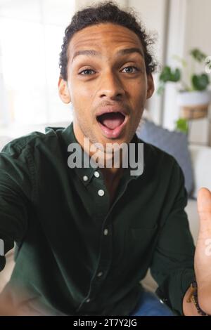 Glücklicher birassischer Mann mit langen lockigen Haaren, der Videoanruf hat und den Mund im sonnigen Wohnzimmer öffnet Stockfoto