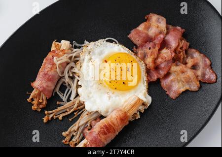 Enoki-Brötchen mit gebratenem Schinken und Spiegelei. Nahaufnahme von goldenem Nadelpilz mit geschnittenem Schinken auf einem schwarzen Teller. Draufsicht, flach. Stockfoto