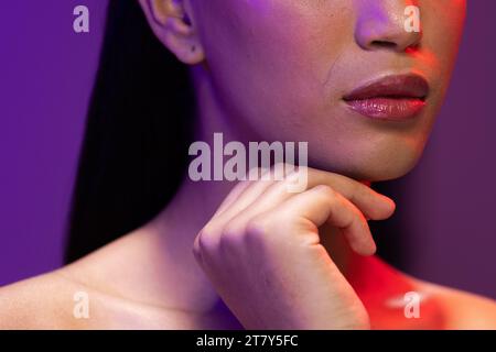 Nahaufnahme einer zweirassigen Frau mit dunklem Haar, roten Lippen und Hand-zu-Gesicht auf Neonhintergrund Stockfoto