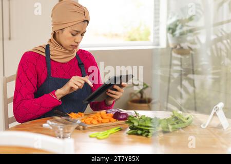 Birassische Frau in Hijab und Schürze, die Gemüse mit Tablette in der Küche zerkleinert Stockfoto